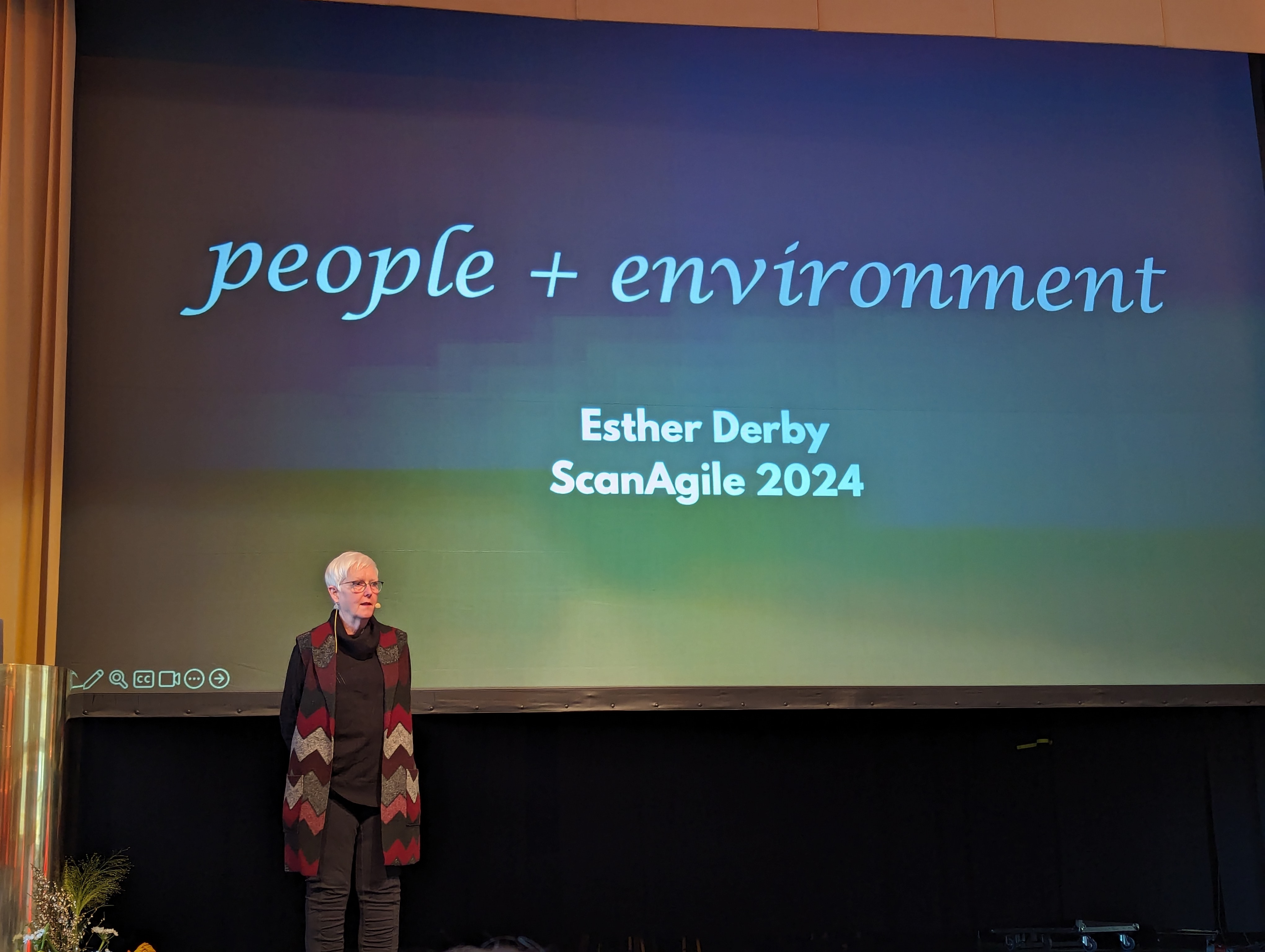 Esther Derby standing on stage with her presentation in the background showing the title &ldquo;people + environment&rdquo;.
